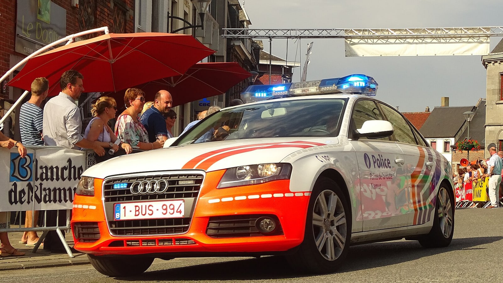 A police car with orange and white livery drives through a street during a public event, passing under red umbrellas as spectators watch… — illustrative photograph