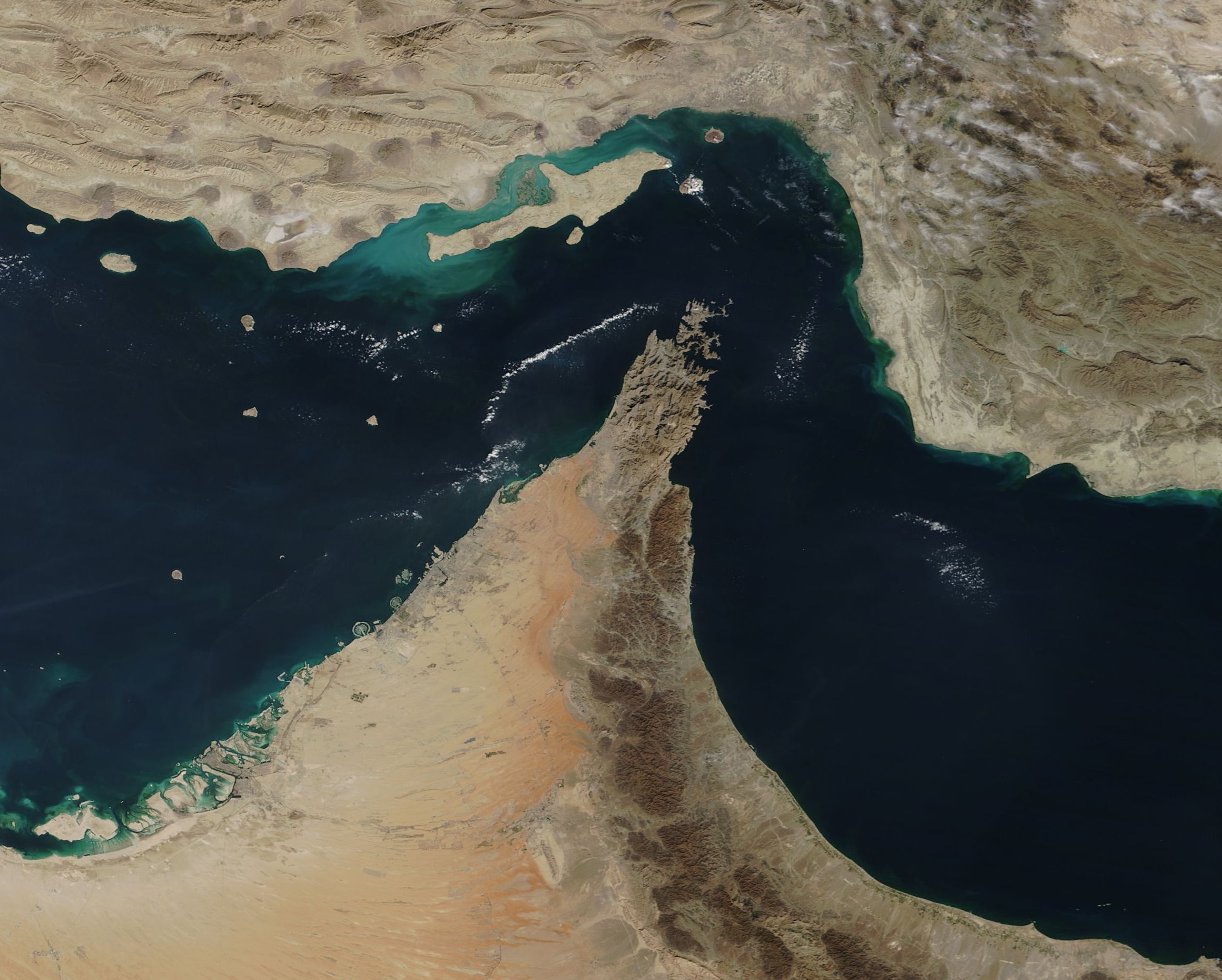 Satellite view of the Strait of Hormuz, showing the Musandam Peninsula and Arabian coastline separated by a narrow channel of dark water from the Iranian coast.